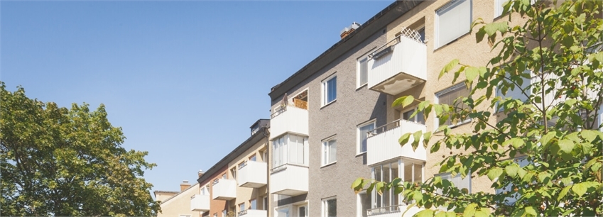 Om projektet - Toppen av en rad fastigheter i gult, brunt och orange med vita balkonger. Blå himmel och grönska.