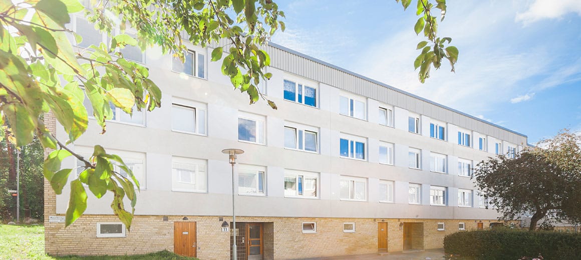 Ljust trevåningshus med grön omgivning.