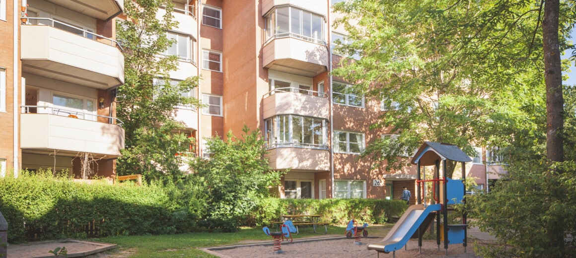 En solig, lummig gård med lekplats framför ett aprikost hus.